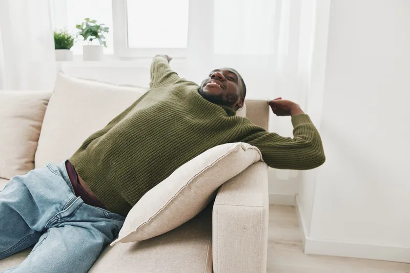 Relaxed young African man enjoying leisure time at home in cozy sweater and jeans, expressing comfort and tranquility in a bright, minimalistic living room