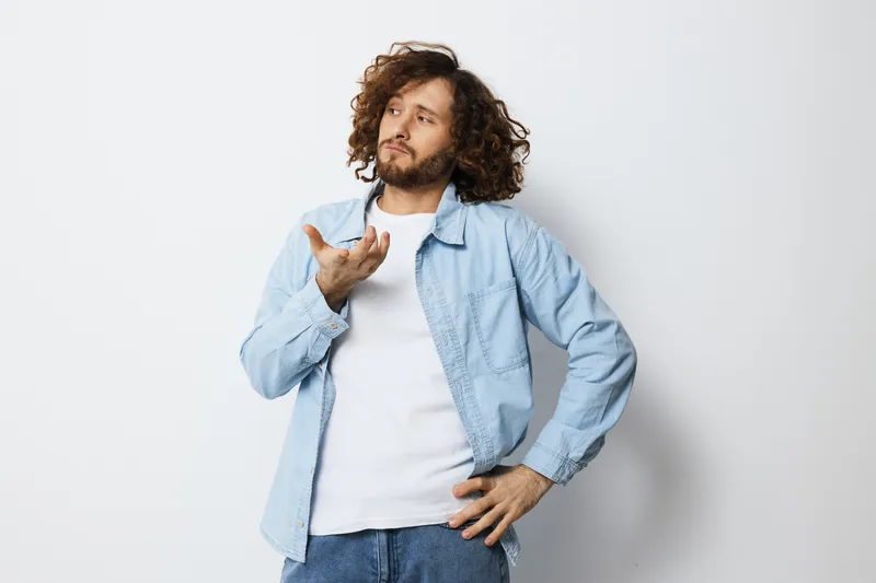 Pensive young man with curly hair in a light blue shirt expressing confusion against a plain white background in casual style