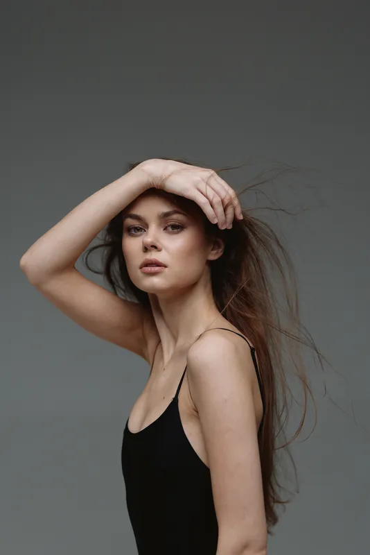 Elegant woman with long hair posing against a gray background wearing a black sleeveless top, showcasing natural beauty and grace.