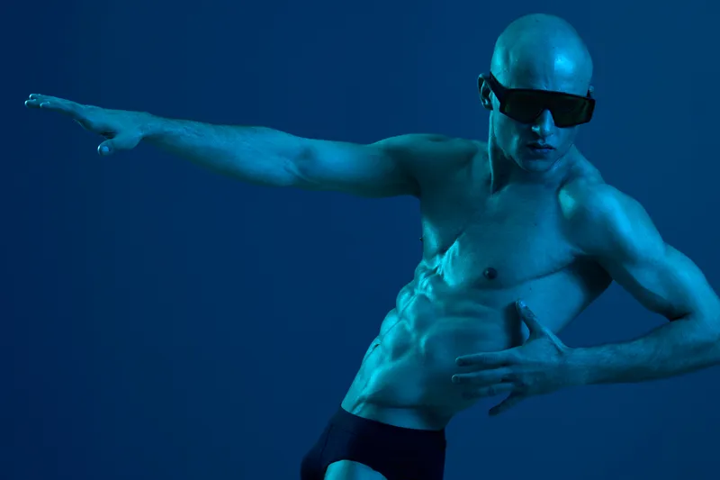 Dynamic pose of a man in dark sunglasses against a blue background, showcasing athleticism and strength in a visually striking manner.