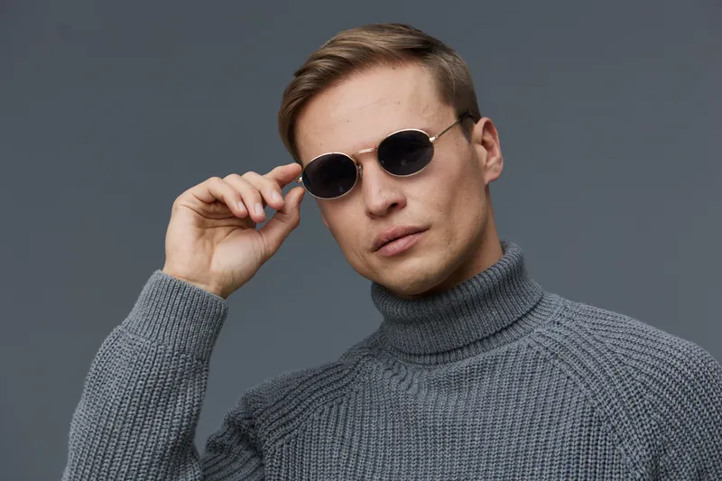 Stylish young man wearing sunglasses and grey turtleneck sweater looking confident isolated on plain grey background posing for camera digital nomad concept