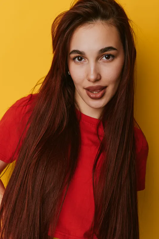 Young woman with long red hair wearing red shirt posing confidently against yellow background, looking directly at camera with slight smile and natural makeup.