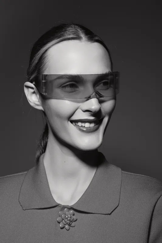 Smiling white woman with light skin wearing transparent sunglasses and a collared shirt with a brooch in a studio with dark background.