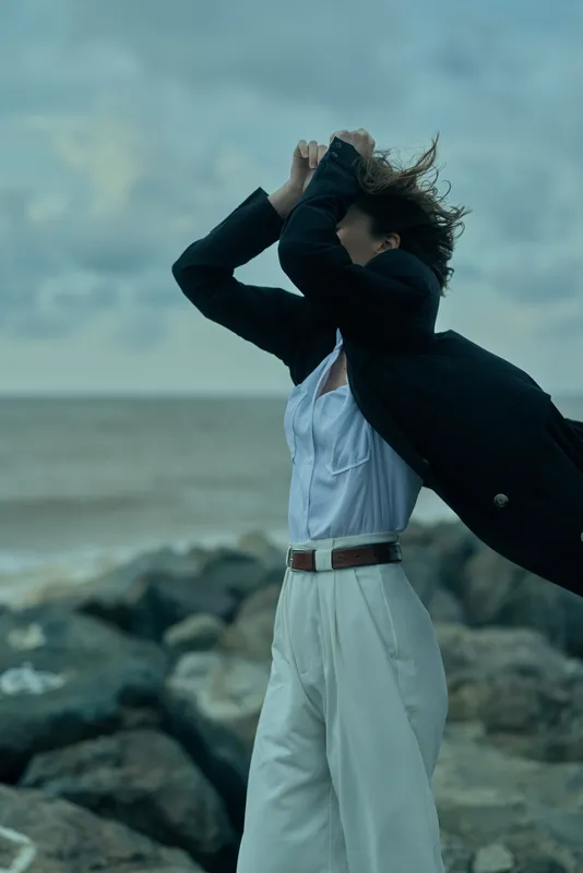 Windy beach scene shows a professional woman in a white suit and dark blazer, arms raised as the coat flares, ocean rocks in the background, conveying motion, freedom, and bold work attitude.