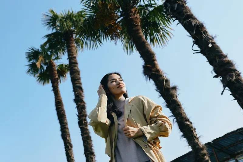 Woman standing beneath tall palm trees in bright sunlight, wearing a light jacket, looking up with a relaxed pose in an outdoor fashion scene.