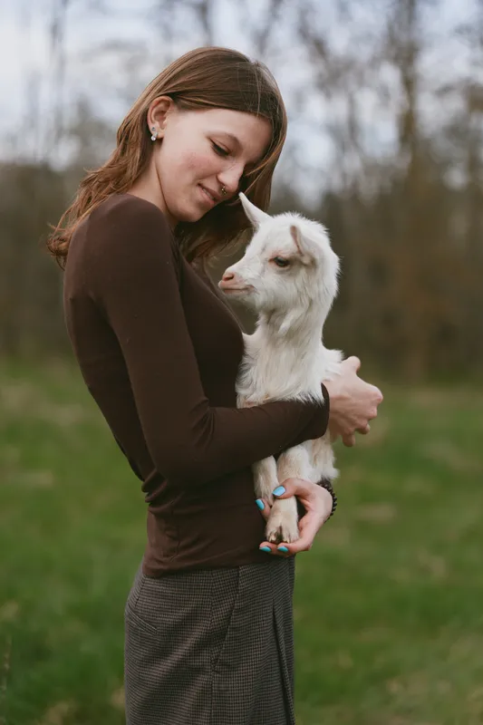Woman with brown sweater hugs a white goat kid in a countryside field, portraying gentle affection, bonding with nature, and calm wholesome lifestyle