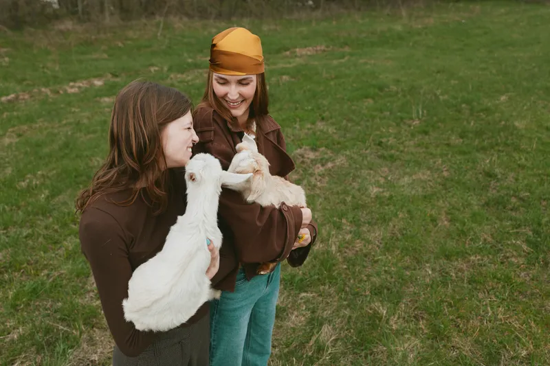 Lifestyle outdoor scene with two women in a grassy field, goat kid held between them, laughter and warmth, casual clothing, natural colors, affection and friendship shown, calm rural moment, joyful