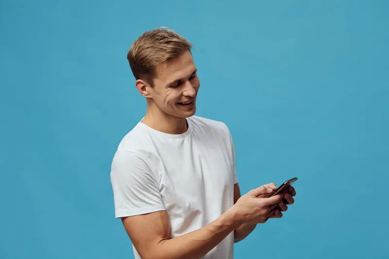 Smiling young man with short brown hair wearing casual white t-shirt, looking at smartphone, standing against plain blue background, isolated, lifestyle concept, modern technology.