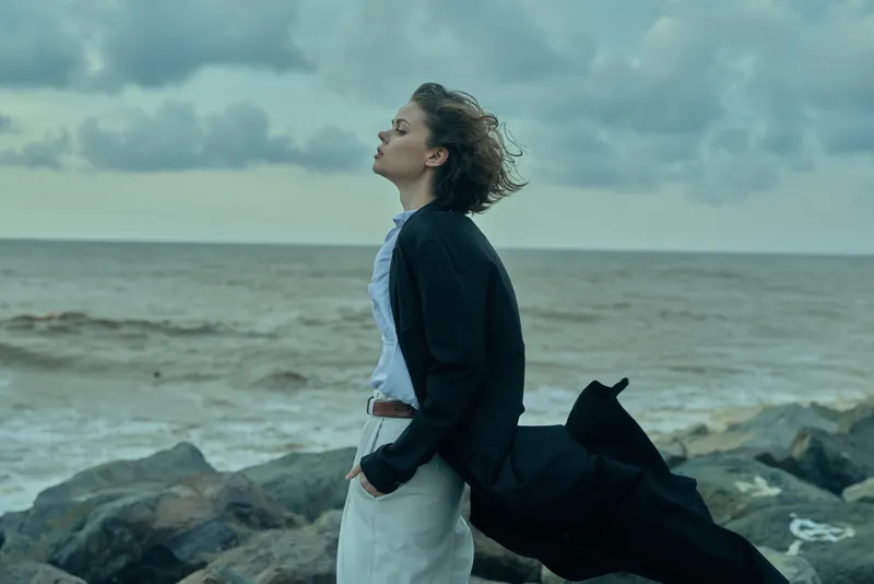 Sea and ocean breeze frame a woman in a blazer and flowing coat on a rocky coast, eyes forward, confident stance against the wind and waves and distant horizon.
