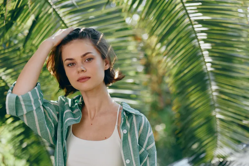 Woman outdoors in natural light, casual outfit, and a calm expression. Green foliage creates a soft backdrop for a relaxed, confident portrait highlighting beauty and everyday mood.