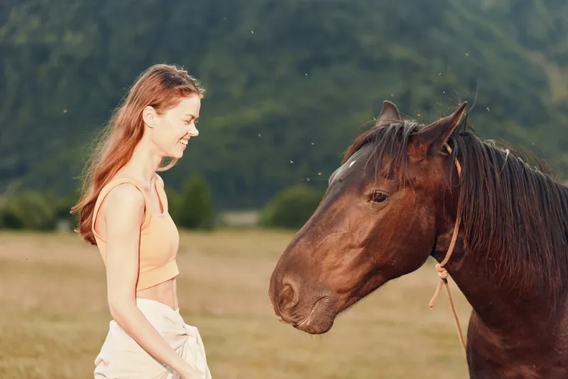 A cheerful woman stands beside a horse in an open field, smiling as they share a calm moment. Natural light and expansive countryside create a tranquil, pastoral scene.