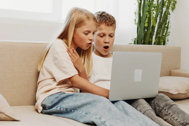 Two surprised children sit on a beige couch looking at a laptop screen with wide eyes. Bright living room with natural light and a green plant in the background creates a cozy atmosphere.