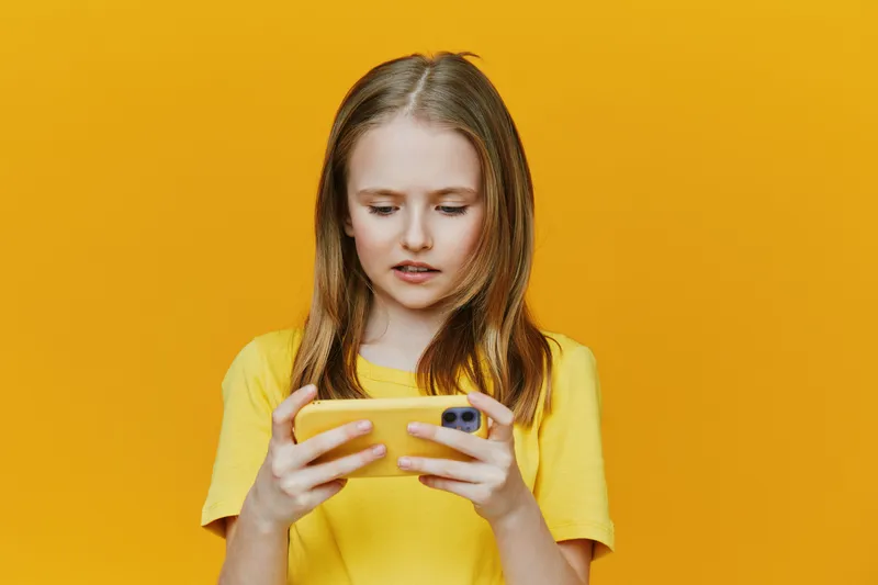 Young girl with straight hair wearing a yellow shirt, focused on her smartphone against a vibrant yellow background, showcasing curiosity and engagement in a playful setting
