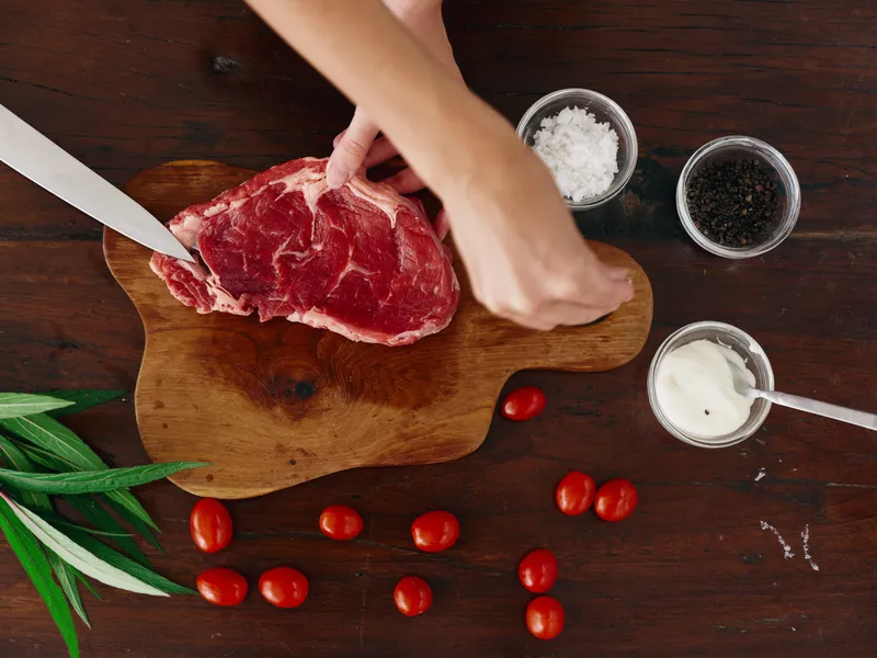 Женщина с ножом в руке нарезает свежее мясо стейка для жарки на кухне, на столе соль, перец и другие специи, красные помидоры черри и зелень, приготовление ужина в домашней кухне