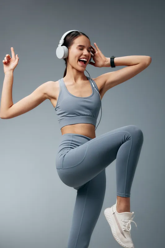 Young woman fitness athlete dancing and exercising in matching grey sports bra and leggings, wearing white headphones and sneakers, dynamic workout against neutral grey background.