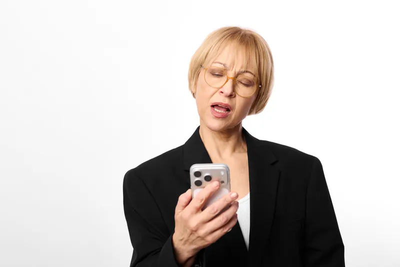 Woman smartphone glasses surprised business suit senior communication technology screen, blonde senior holds mobile phone, shocked face, studio portrait on white background isolated