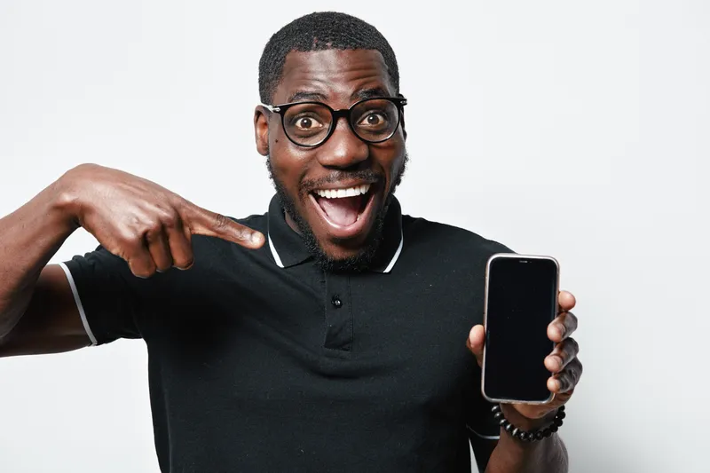 African American man with dark skin and glasses smiling and pointing at smartphone screen in black polo shirt on white background. Excited male showing mobile device.