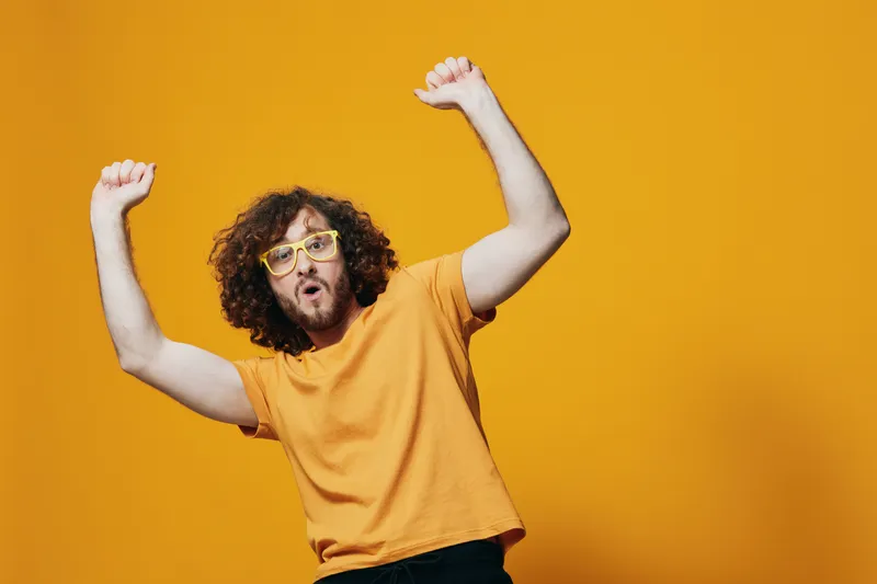 Young man celebrating with arms raised against a vibrant yellow background, showcasing joy, confidence, and playful energy in a casual orange t shirt