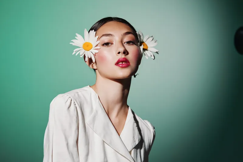 Young woman wearing a cream blazer and daisies in her hair poses confidently against green background. This creative beauty and fashion portrait highlights natural elegance.