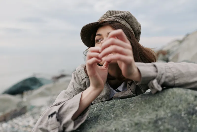 A thoughtful young woman wearing a tweed cap leans on a large rock outdoors, gazing into the distance. Moody natural light and casual clothing create a contemplative atmosphere.