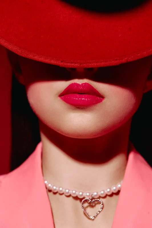 Close-up of young woman wearing a large red hat contrasting with pink blazer, bright red lipstick lips visible, delicate pearl necklace with heart pendant. Creative fashion and beauty concept.