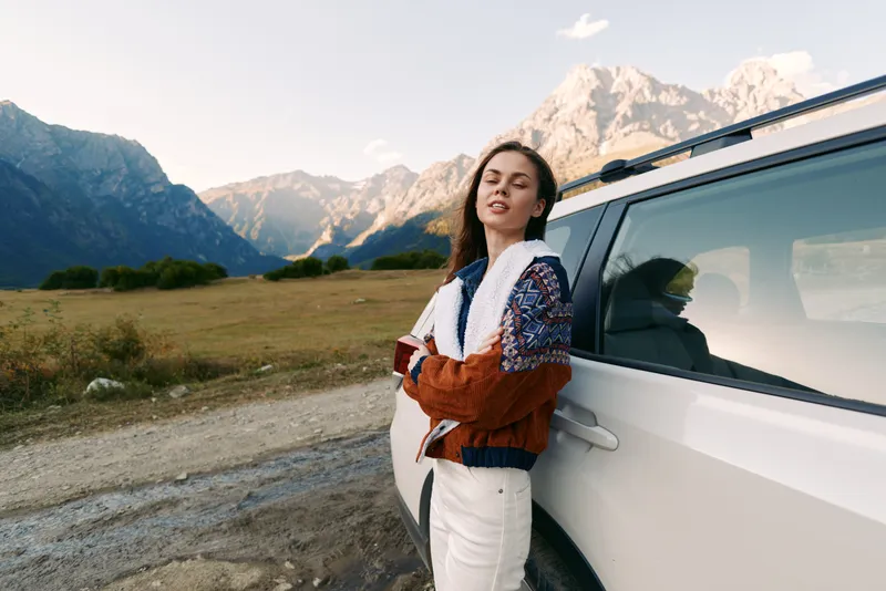 Woman leaning against white car in the mountains, travel portrait with passport and casual jacket, outdoor road trip scene capturing scenic peaks, meadow and relaxed mood.