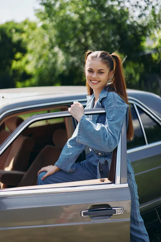 Joyful young woman with ponytails in denim outfit posing by vintage car, showcasing a cheerful expression against a lush green background.