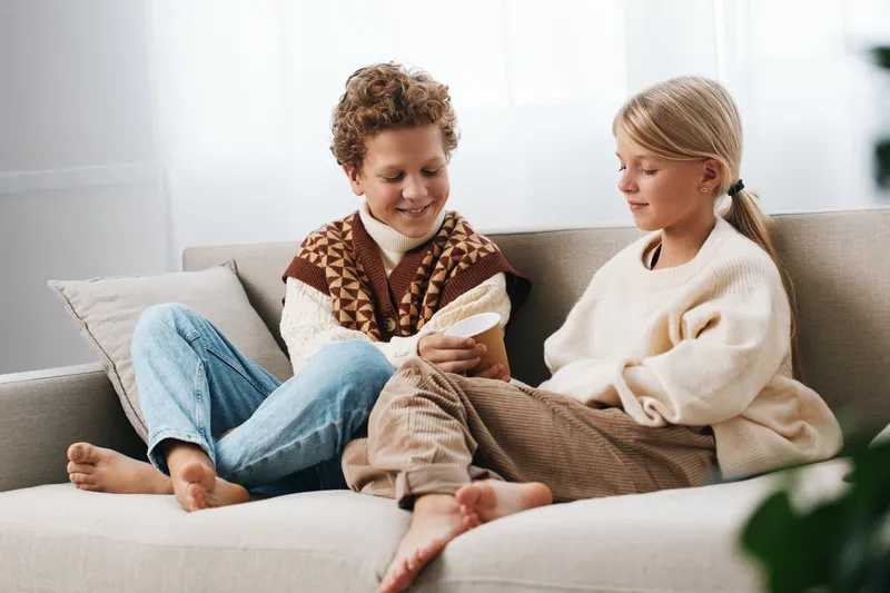 Two children sit on a cozy sofa, sharing a small device and smiling as they explore a screen together in a bright living room, enjoying a calm home moment.