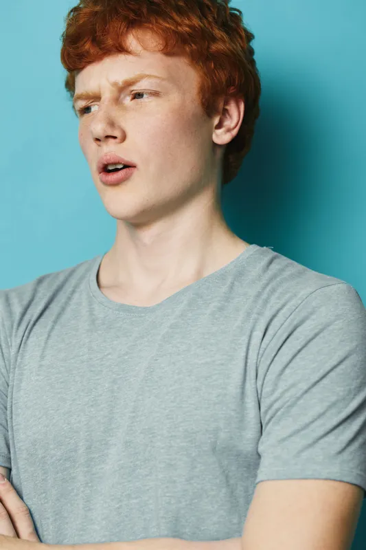 Young man with red hair wearing gray t shirt standing against blue background with confused expression looking aside. Casual style, portrait and emotions, isolated studio shot.