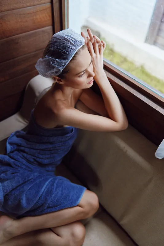 young woman relaxing in sauna wrapped in blue towel with hair cap, sitting by window enjoying peaceful moment and warm wooden interior