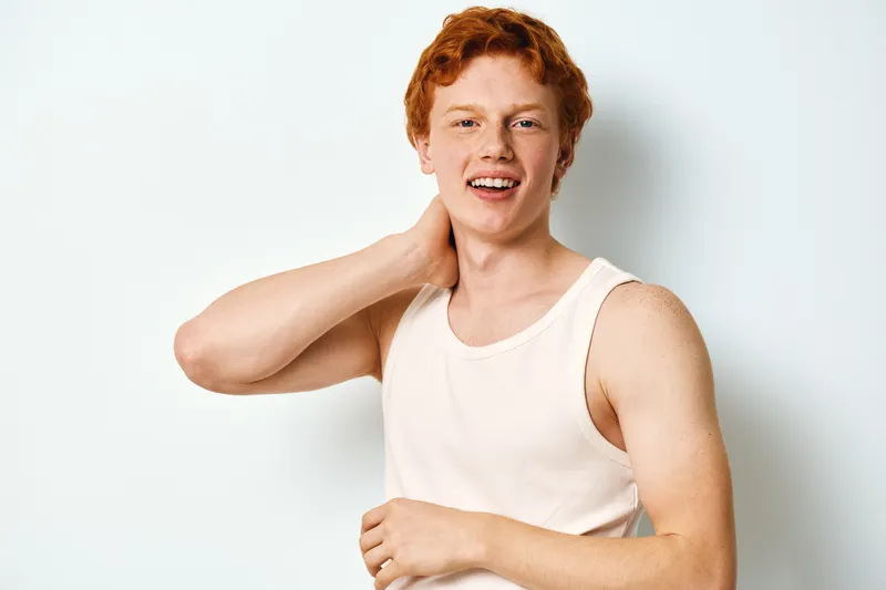 Young man with red hair and fair skin posing in white sleeveless shirt against white background with confident smile and hand touching neck.