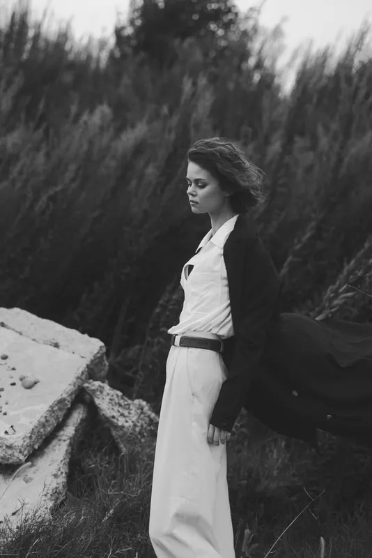 Woman in a long white dress and blazer stands near rugged rocks as wind tousles hair and cape, monochrome outdoor fashion. A moody, cinematic moment highlighting movement and style in nature