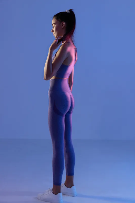 Woman sport fitness wearing purple workout clothes and white sneakers, standing in profile with hands touching neck, illuminated by vibrant blue and pink lighting.