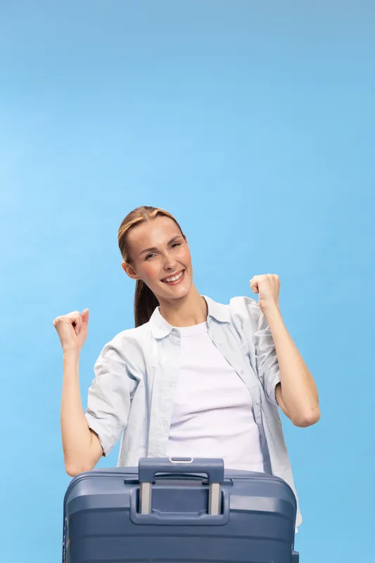 Happy young woman with suitcase celebrates travel plans on blue background. Casual outfit and joyful expression show excitement for vacation and adventure. Concept for tourism and journey.