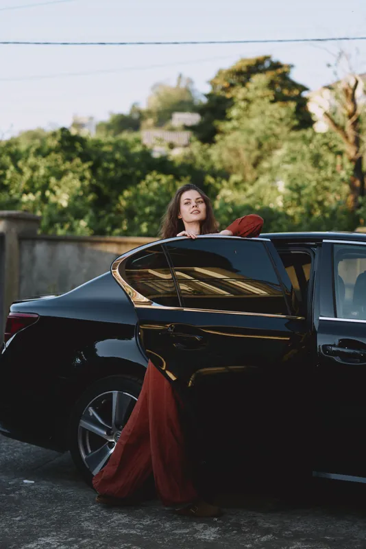 Woman lifestyle portrait as she emerges from a sleek car wearing a flowing red dress, captured in a sunny outdoor setting with greenery and a confident glamorous mood