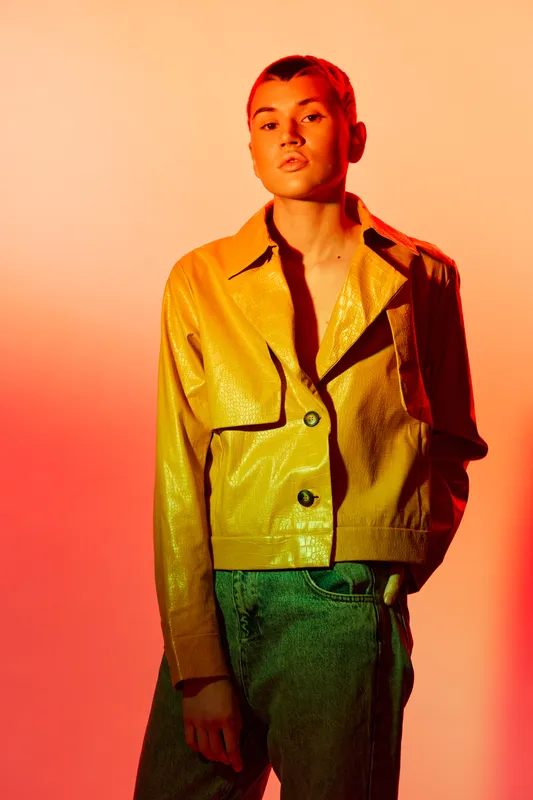 Young woman with short hair wearing a shiny yellow jacket and green jeans, posing confidently against a warm orange gradient background in stylish fashion.