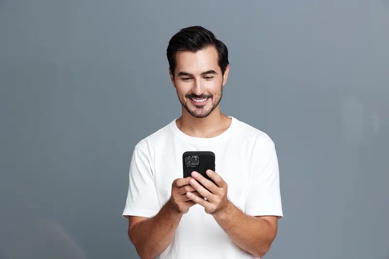 Smiling man wearing white t-shirt looking at smartphone with happy expression while standing against plain grey background. Lifestyle and technology concept.