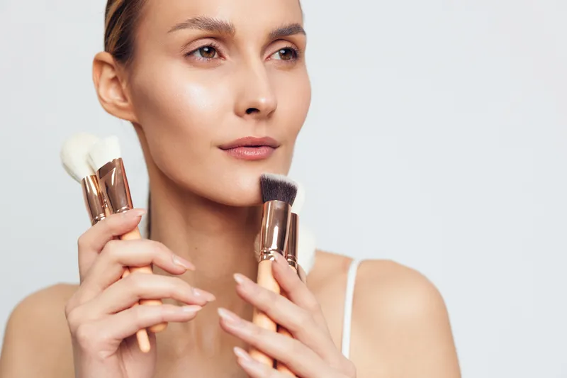 Young woman holding makeup brushes near her face with neutral expression. Studio portrait on white background. Concept of beauty, cosmetics, skincare, and professional makeup tools.