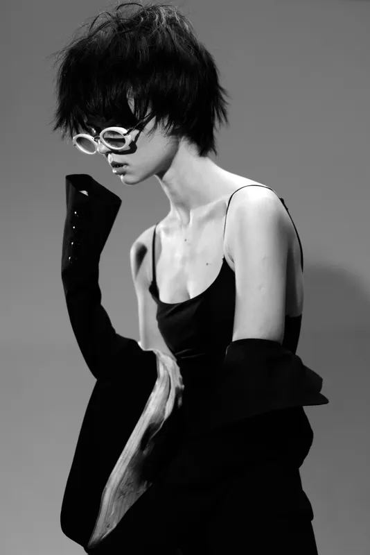 Black and white portrait of a stylish woman wearing white sunglasses and dark clothing with edgy fashion and moody expression.