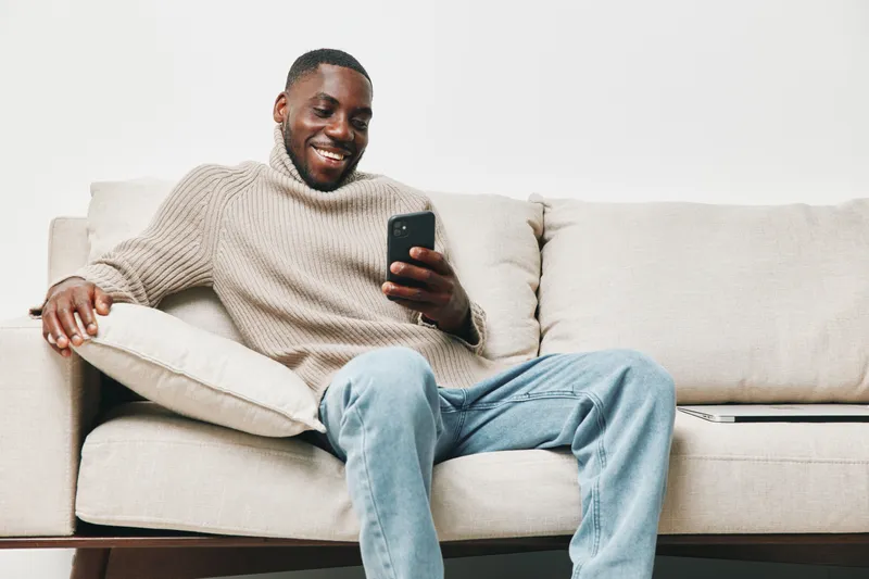 Smiling Black man in a cozy sweater using smartphone on a comfortable couch, showcasing a relaxed atmosphere and modern lifestyle