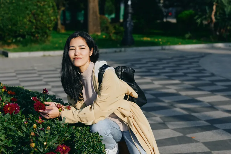 Woman with backpack crouching beside blooming flowers in a park, enjoying a sunny day, casual attire, candid outdoor moment focused on nature, relaxation and strolls.
