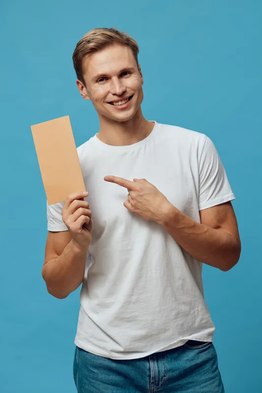 Smiling young man holding a blank cardboard sign pointing to it with his finger, wearing casual white t-shirt and jeans, standing against a bright blue background in a studio setting. People lifestyle