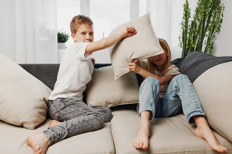 Two playful children sitting on a beige couch having a pillow fight in a bright living room. The boy and girl enjoy a fun moment with casual clothes and relaxed barefoot posture.