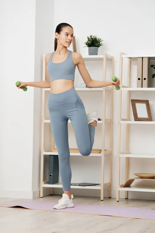 Woman in athletic wear performs a balance drill with dumbbells at home, indoor gym backdrop with shelves and a plant, showcasing focused fitness and strength training.