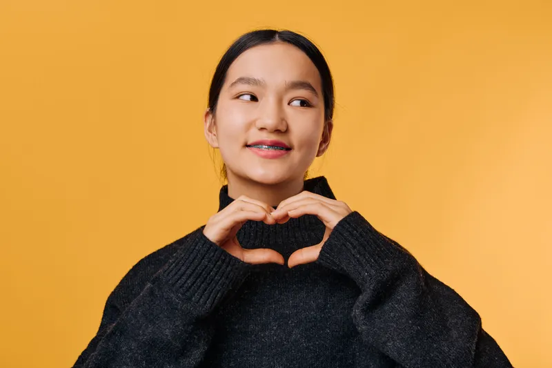 Young woman in black sweater with braces making a heart shape with her hands, smiling and looking sideways on yellow background for love and positive emotions.