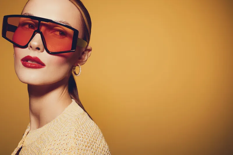 Stylish woman with oversized orange sunglasses and red lipstick poses against a warm yellow background. Fashion concept with bold accessories and confident expression.