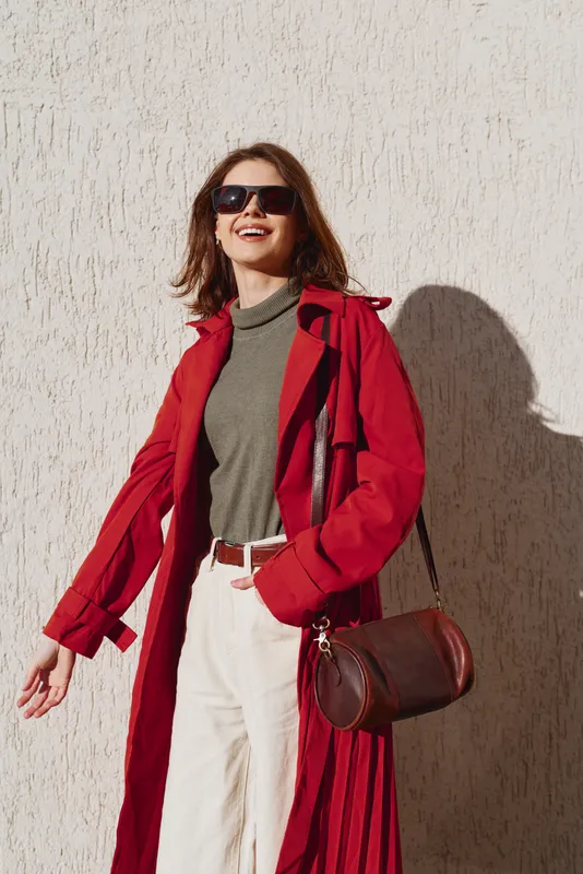 Smiling woman wearing sunglasses and red coat standing against white wall, casual outfit with brown handbag, bright sunlight casting strong shadow