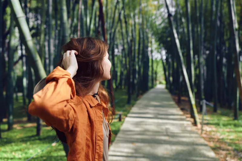 Woman on a path in a bamboo forest wearing an orange jacket and hat, walking and looking back along a sunlit trail, nature escape and peaceful outdoor portrait.