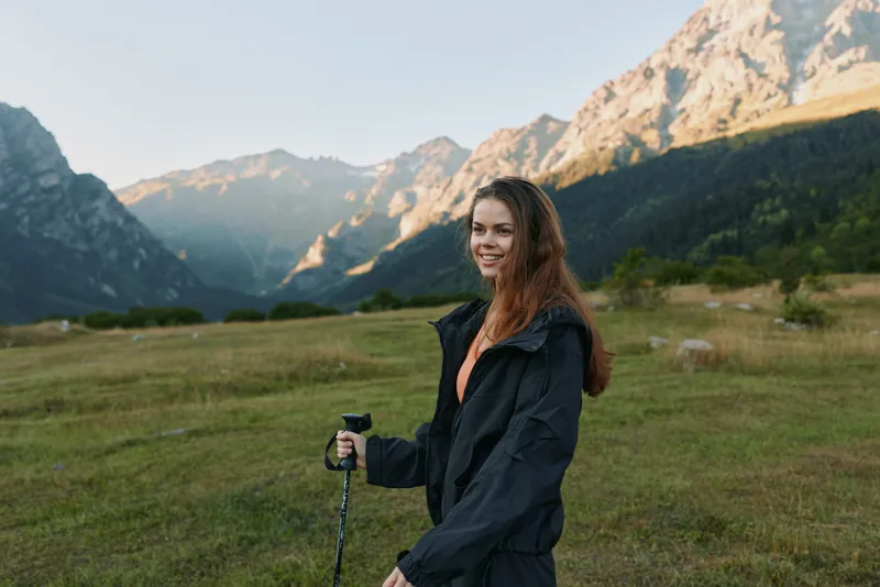 A cheerful woman hiker stands in a wide alpine meadow with mountains in the distance. She wears a dark jacket and grips a trekking pole, smiling as sunlight highlights the scene, conveying outdoor