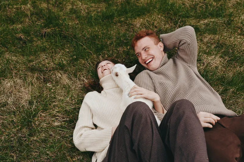 A joyful couple lounge on grass with a white goat kid, wrapped in cozy sweaters, enjoying a carefree countryside moment. A lifestyle scene of connection, playfulness, and rural bonding.